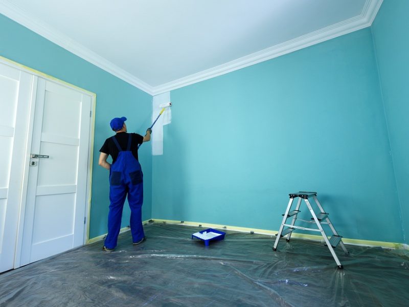 Man in a working overall is painting the wall in white color